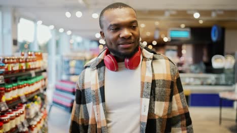 A-happy-and-confident-man-with-Black-brunette-skin-color-in-a-checkered-shirt-and-red-wireless-headphones-walks-along-the-counter-in-a-modern-spacious-and-sunny-supermarket-and-looks-at-the-goods-on-display