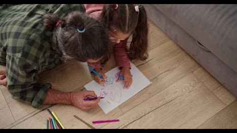 Top-view-of-a-little-brunette-girl-in-a-pink-dress-draws-on-a-white-sheet-of-paper-together-with-her-lonely-father,-a-brunette-man-in-a-Green-checkered-shirt,-using-multi-colored-markers-while-lying-on-the-floor-near-a-gray-sofa-in-a-modern-apartment