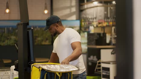 A-delivery-man-with-Black-skin-in-a-white-T-shirt-happy-and-joyful-puts-all-the-necessary-purchased-products-into-a-yellow-bag-while-standing-at-the-checkout.-Happy-male-delivery-man-buys-the-necessary-goods-and-puts-them-in-his-bag-to-deliver-to-the-client