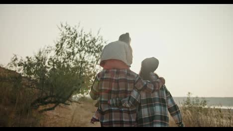 Rear-view-of-a-happy-family,-a-brunette-girl-in-a-hat-in-a-green-checkered-shirt-walks-with-her-husband,-a-man-in-a-brown-checkered-shirt,-and-their-blonde-daughter-in-a-white-jacket-sits-on-the-man’s-shoulders-during-their-hike-and-walk-outside-the-city