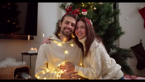 Portrait-of-a-happy-couple,-a-brunette-guy-and-a-girl-in-a-white-sweater-and-in-New-Year's-hoops-with-a-smile,-holding-hands-and-hugging-each-other,-decorated-with-a-New-Year's-glowing-garland-near-a-green-Christmas-tree-in-a-cozy-room-in-a-winter-evening