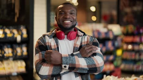 Portrait-of-a-happy-and-confident-man-with-Black-skin-color-with-a-beard-in-a-checkered-shirt-and-red-wireless-headphones-who-crosses-his-arms-on-his-chest-miles-and-poses-among-the-counters-in-a-modern-grocery-store