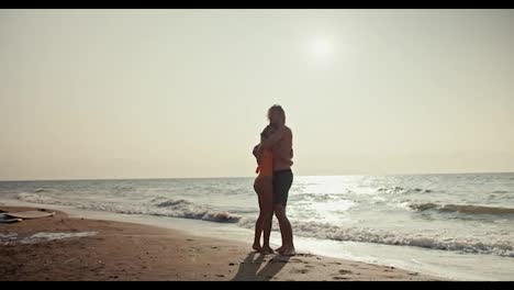 Happy-couple,-a-blond-guy-in-shorts-and-a-blonde-girl-in-an-orange-swimsuit-are-hugging-while-standing-on-a-brown-rocky-seashore-in-the-morning