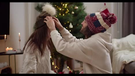 A-happy-guy-in-a-New-Year's-warm-hat-puts-a-green-brown-hat-on-the-head-of-his-brunette-girlfriend-in-a-White-sweater.-They-look-at-each-other-and-smile-near-the-New-Year-tree-in-the-atmosphere-of-Christmas-and-New-Year-at-home-in-the-evening