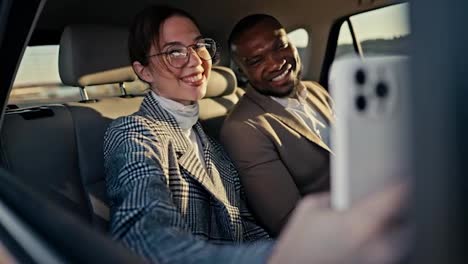 A-happy-brunette-girl-in-round-glasses-takes-a-selfie-with-her-colleague-a-Black-skinned-businessman-in-a-brown-suit-during-their-shared-trip-in-the-interior-of-a-modern-car