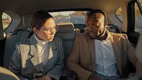 Confident-man-Businessman-with-Black-skin-color-in-a-brown-suit-talks-about-his-ideas-and-plans-to-his-colleague-a-brunette-girl-in-round-glasses-and-a-gray-jacket-during-his-trip-in-a-modern-car-outside-an-urban-area