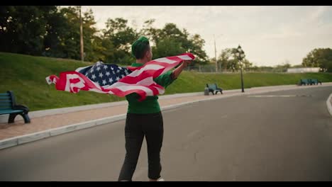 Una-Niña-De-Pelo-Corto-Verde-Con-Una-Camiseta-Verde-Y-Pantalones-Negros-Corre-Sosteniendo-Detrás-De-Sus-Hombros-La-Bandera-De-Los-Estados-Unidos-De-América,-Que-Ondea-Con-El-Viento-En-El-Parque-En-Verano.