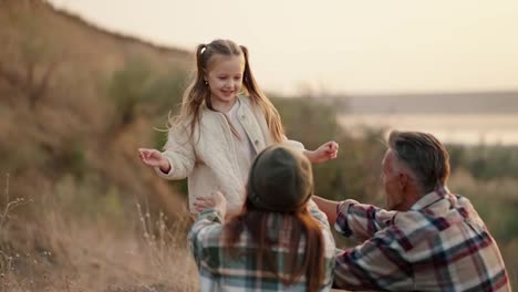 A-happy-little-blonde-girl-in-a-white-jacket-runs-to-her-parents,-jumps-on-them-and-they-fall-on-the-mat-during-their-outdoor-recreation-and-picnic-outside-the-city-in-the-summer.-Happy-girl-runs-to-hug-her-parents-during-a-family-picnic