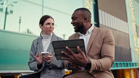 A-brunette-girl-in-round-glasses-and-a-gray-jacket-communicates-with-a-man-with-Black-skin-in-a-brown-jacket-with-his-businessman-colleague-during-their-common-plans-on-a-Black-Tablet-near-a-large-building