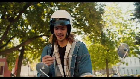 A-guy-with-curly-hair-in-a-white-moped-helmet-in-a-denim-shirt-sits-on-a-moped-and-smokes-in-a-summer-city-on-the-street.-The-guy-took-a-break-while-riding-a-moped-through-the-city-streets-in-the-summer