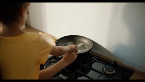 Rear-view-of-a-little-brunette-girl-in-a-yellow-dress-together-with-her-mother-breaking-a-chicken-egg-into-a-frying-pan-and-preparing-fried-eggs-together-in-a-modern-kitchen