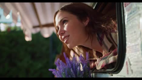 Happy-brunette-girl-in-a-plaid-shirt-sticks-her-head-out-to-the-city-and-looks-around-from-a-trailer-during-her-picnic-at-a-camp-outside-the-city-in-summer.-Happy-brunette-girl-looking-out-of-trailer-in-summer