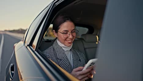 Außenansicht-Eines-Glücklichen-Brünetten-Mädchens-In-Runder-Brille,-Geschäftsfrau-Kommuniziert-Auf-Einem-Weißen-Telefon-Und-Schaut-Aus-Dem-Fenster-Während-Ihrer-Fahrt-In-Einem-Modernen-Auto