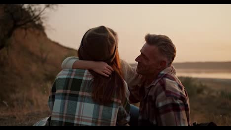 Primer-Plano-De-Una-Niña-Rubia-Con-Una-Chaqueta-Blanca-Que-Corre-Hacia-Sus-Padres,-Que-Están-Sentados-En-Una-Colchoneta-Durante-Un-Picnic-Fuera-De-La-Ciudad-Por-La-Tarde-En-Verano.-Niña-Feliz-Corre-Hacia-Sus-Padres-Durante-Un-Picnic-Fuera-De-La-Ciudad-En-Verano.