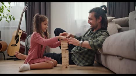 A-lonely-father,-a-brunette-man-in-a-Green-checkered-shirt-plays-with-his-little-daughter,-a-brunette-girl-in-a-pink-dress,-in-the-board-game-Jenga,-leaning-on-a-gray-sofa-on-the-floor-in-a-modern-apartment