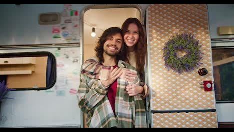 Portrait:-Happy-brunette-girl-hugs-her-brunette-boyfriend-with-stubble-in-a-Green-checkered-shirt-and-a-red-T-shirt-while-standing-near-her-trailer-during-her-picnic-outside-the-city-in-the-summer-at-the-camp