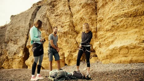 A-blonde-girl-in-a-blue-jacket,-a-blonde-girl-with-curly-hair-in-black-clothes-and-a-brunette-guy-in-a-gray-T-shirt-put-on-a-belay-before-they-start-rock-climbing-on-a-rocky-shore-near-the-yellow-rocks-during-the-day