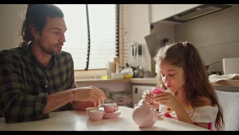 Happy-brunette-man-in-green-checkered-shirt.-Lonely-father-plays-tea-party-game-with-his-little-daughter-brunette-girl-in-pink-dress-using-toy-pink-cups-and-kettle-in-kitchen