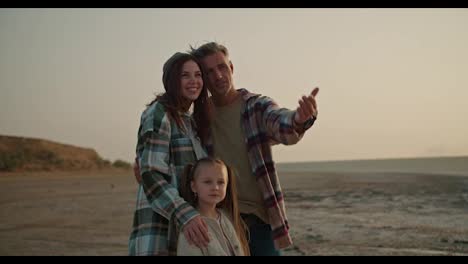 Happy-brunette-man-with-gray-hair-in-a-checkered-shirt-stands-and-enjoys-the-view-with-his-brunette-wife-in-a-green-checkered-shirt-and-little-blonde-daughter-during-his-vacation-outside-the-city-on-a-deserted-seashore-in-the-evening-in-summer