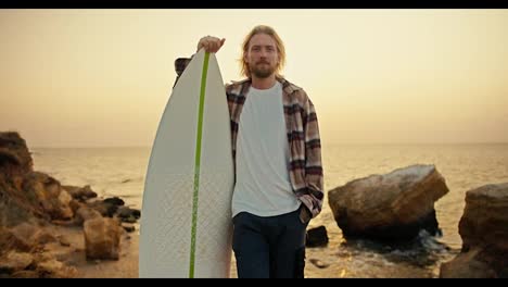 Portrait-of-a-tall-blond-guy-with-a-beard-in-a-plaid-shirt-and-a-white-T-shirt-who-stands-and-holds-his-white-surfboard-near-him-on-a-rocky-shore-near-the-sea-early-in-the-morning-at-Sunrise