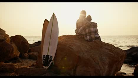 Rear-view-of-a-blond-guy-in-a-plaid-shirt-and-his-girlfriend-in-a-black-hat,-turning-on-the-shirt,-sitting-and-hugging-on-the-Big-Stone-on-a-rocky-shore-by-the-sea,-near-them-there-are-their-white-and-yellow-surfboards-at-Sunrise-in-the-morning