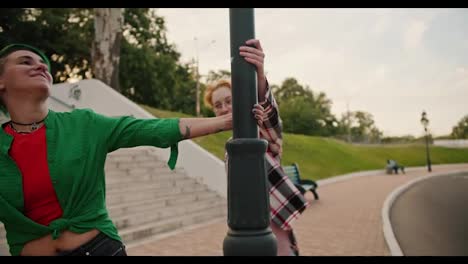 Two-lesbian-girls-with-short-haircuts-of-bright-colors-in-pink-and-green-checkered-shirts,-during-their-date,-spin-around-a-lamppost-and-continue-to-walk-further-holding-hands-in-the-park