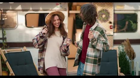 A-happy-brunette-guy-with-a-green-checkered-shirt-dances-A-cheerful-dance-with-his-brunette-girlfriend-in-a-light-brown-shirt-near-their-trailer-in-the-camp-during-a-picnic-and-vacation-outside-the-city-in-the-summer