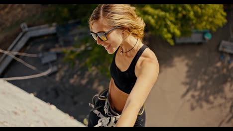 Portrait-of-a-happy-blonde-girl-in-a-black-sports-summer-uniform-and-in-sunglasses-who-climbed-up-a-white-climbing-wall-and-with-multi-colored-ledges-and-poses-at-a-height-using-a-belay-in-the-summer