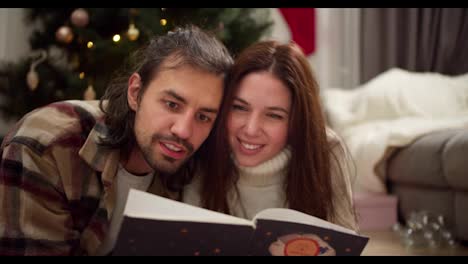 An-interested-guy-in-a-checkered-shirt-and-a-brunette-girl-in-a-white-sweater-with-a-cup-of-hot-drink-in-their-hands-are-reading-a-New-Year's-book-in-the-cozy-atmosphere-of-a-room-decorated-in-New-Year's-style