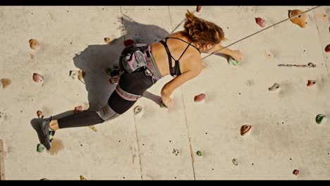 Vertical-video-of-a-blonde-girl-with-a-bob-hairstyle-in-a-black-top-and-a-special-uniform-and-equipment-for-folding-using-belay-climbing-up-the-route-of-a-climbing-wall-in-the-summer