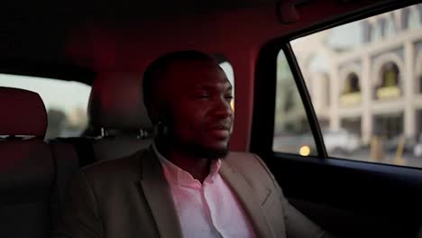 A-happy-man-with-Black-skin-and-a-beard-a-businessman-in-a-brown-suit-rides-in-a-modern-car-interior-around-the-city-during-his-business-trip