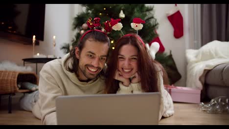 Happy-couple-guy-and-girl-in-New-Year's-clothes-lie-on-the-floor-in-front-of-a-gray-laptop-watching-a-Christmas-movie-near-the-Christmas-tree-in-a-cozy-decorated-room