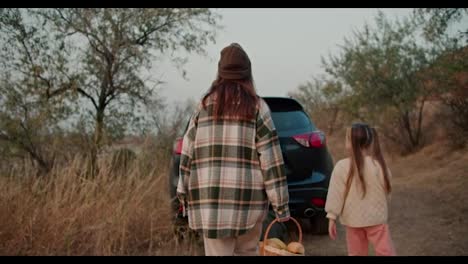 Rear-view-of-a-brunette-girl-in-a-hat-in-a-green-checkered-shirt,-together-with-her-middle-aged-husband-in-a-checkered-shirt-and-little-daughter,-go-to-a-black-car-and-put-their-things-there-after-a-picnic-in-a-summer-evening-outside-the-city