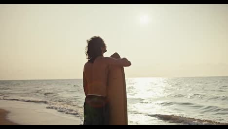 Rear-view-of-a-guy-with-medium-length-hair-with-a-bare-torso-in-shorts-stands-and-holds-his-wooden-yellow-surfboard-on-a-sandy-rocky-shore-near-the-sea-at-Sunrise-in-the-morning