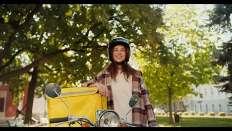 Portrait-of-a-happy-brunette-courier-girl-in-a-checkered-shirt-wearing-a-White-motorcycle-helmet-with-a-yellow-bag-for-food-delivery-on-the-street-in-summer