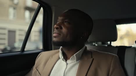 Confident-man-Businessman-with-Black-skin-in-a-brown-suit-rides-in-a-modern-car-and-mysteriously-looks-out-the-window-at-the-houses-and-people-in-the-city