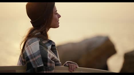 A-happy-blonde-girl-in-a-black-hat-and-a-plaid-shirt-stands-near-a-rocky-shore,-holds-a-surfboard-and-looks-towards-the-sea-in-the-morning