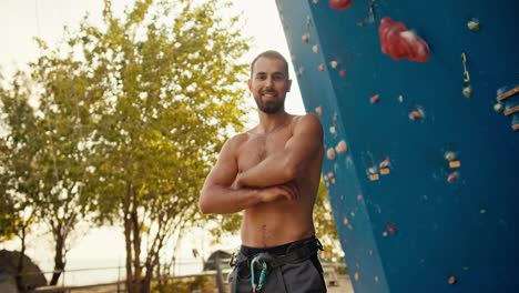 Portrait-of-a-happy-brunette-guy-with-a-beard-who-folds-his-arms-on-his-chest-and-looks-at-the-camera-near-a-blue-climbing-wall-on-a-sunny-summer-day.-Experienced-man-Scala-with-bare-torso-posing-and-looking-at-camera-Near-his-climbing-wall-in-summer