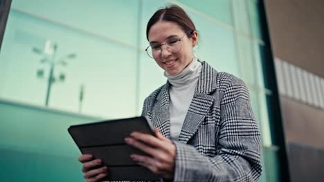 Close-up-of-a-happy-girl-businesswoman-in-round-glasses-in-a-striped-gray-jacket-works-and-looks-into-a-black-tablet-and-also-at-the-street-around-during-her-break-at-work-in-the-city