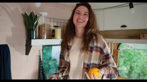 Happy-brunette-girl-in-a-plaid-shirt-juggles-two-oranges-and-dances-in-her-motorhome-or-trailer-outside-the-city-in-the-summer-at-a-camp-on-a-picnic.-Happy-girl-juggling-fruits-and-having-fun-in-her-house-in-summer