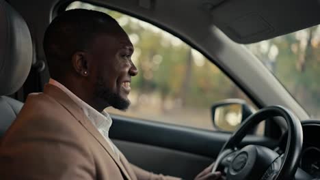 Un-Hombre-De-Negocios-Feliz-Y-Sonriente,-De-Piel-Negra-Y-Barba-Con-Una-Chaqueta-Marrón,-Conduce-Un-Automóvil-Moderno-Durante-Su-Viaje-De-Negocios-Por-La-Ciudad