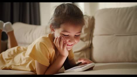 Close-up-shot:-Portrait-of-a-happy-little-brunette-girl-lying-on-a-light-brown-sofa-looking-at-something-on-her-phone-smiling-and-looking-at-the-camera-in-a-modern-room