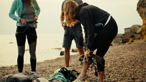 A-girl-climber-puts-on-a-special-belay-and-attaches-a-special-rope-to-her-belt-in-order-to-start-rock-climbing-on-a-rocky-shore-near-the-rocks-by-the-sea.-A-group-of-rock-climbers-put-on-their-gear-and-prepare-to-climb-the-rocks