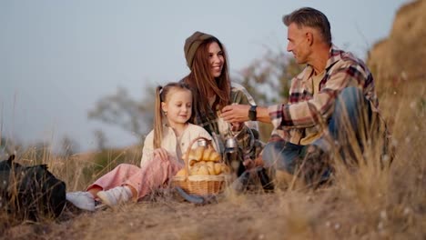 Plano-Lateral-De-Un-Hombre-Feliz-De-Mediana-Edad-Con-Cabello-Gris-Y-Una-Camisa-A-Cuadros-Que-Sirve-Té-Para-Su-Esposa-Y-Su-Pequeña-Hija-Durante-Su-Picnic-Fuera-De-La-Ciudad-En-La-Tarde-De-Verano.