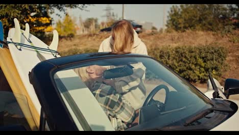 A-blonde-girl-in-a-white-sweatshirt-communicates-with-her-blonde-boyfriend-with-a-beard-in-a-checkered-shirt-who-is-sitting-in-a-White-convertible-with-surfboards-in-the-city