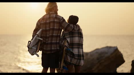 Rear-view-of-a-happy-couple,-a-blond-guy-with-a-beard-in-a-checkered-shirt-stands-with-his-blonde-girlfriend-in-a-black-hat-and-a-checkered-shirt,-they-hold-surfboards-in-their-hands-and-look-towards-the-sea-in-the-morning-at-sunrise