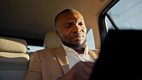 Confident-and-thoughtful-man-Businessman-with-Black-skin-color-in-a-brown-suit-reviews-his-plans-and-draws-with-his-finger-on-a-tablet-while-driving-in-a-modern-salon-in-a-car-in-an-urban-area