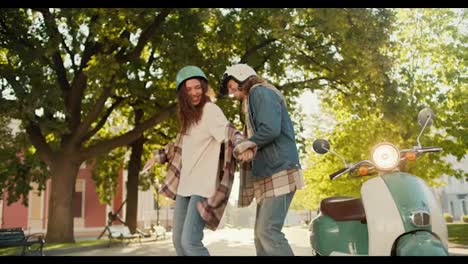 Happy-girl-in-a-checkered-shirt-and-a-motorcycle-helmet-in-denim-pants-dances-with-her-boyfriend-in-a-denim-jacket-near-her-green-moped-in-a-sunny-summer-park-in-the-city