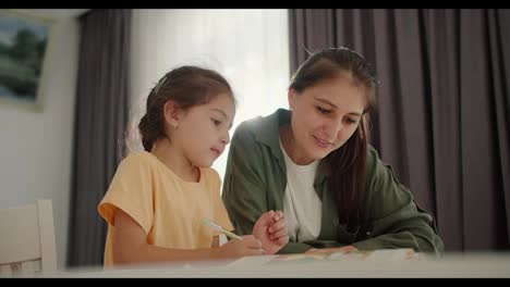 A-little-brunette-girl-in-a-yellow-dress-does-her-homework-with-her-mother,-a-brunette-woman-in-a-green-jacket,-and-writes-in-her-notebook-while-sitting-at-the-table-in-a-cozy-modern-apartment