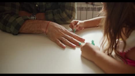 Close-up-shot-of-a-little-brunette-girl-in-a-pink-dress-doing-a-manicure-to-her-dad,-a-man-in-a-green-checkered-shirt,-using-green-polish-on-the-kitchen-table-at-home